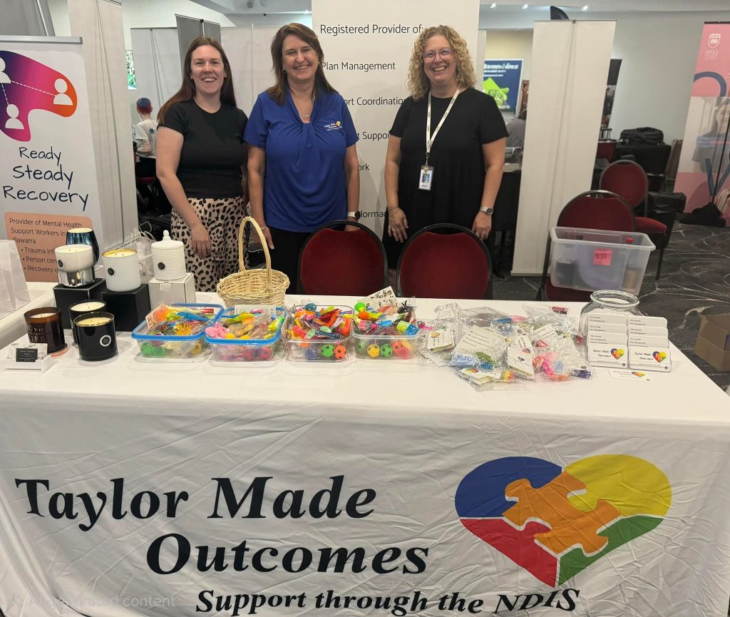 TMO-expo Three Taylor Made Outcomes team members at their stall at a community expo, with a TMO banner and sensory items laid out on the table.